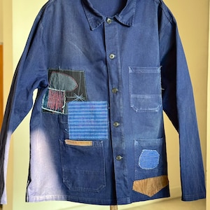 May include: A navy blue jacket with a collared neckline and button closure. The jacket features several patches in various colors and textures, including blue, black, and brown. Two front pockets are visible. The jacket is hanging on a black hanger.