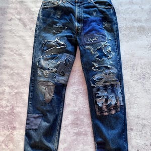 Può includere: Jeans blu con dettagli distressed e toppe. I jeans presentano diverse toppe di varie tonalità di blu e grigio, con strappi e lacerazioni visibili sulle gambe, creando un'estetica unica e usurata.