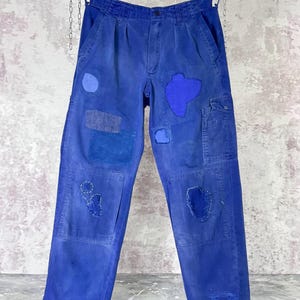Peut inclure: Un pantalon bleu orné de multiples pièces de tissu dans des nuances de bleu variées, incluant des formes circulaires et irrégulières. Le pantalon présente des coutures blanches visibles autour des pièces, une fermeture à bouton et des poches latérales.