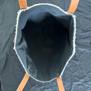 Pu&ograve; includere: Vista aperta di una borsa tote in denim con manici in pelle marrone. L'interno della borsa &egrave; nero e i bordi in denim sono sfilacciati. La borsa ha rivetti in ottone e una costruzione robusta, adatta al trasporto di vari oggetti.