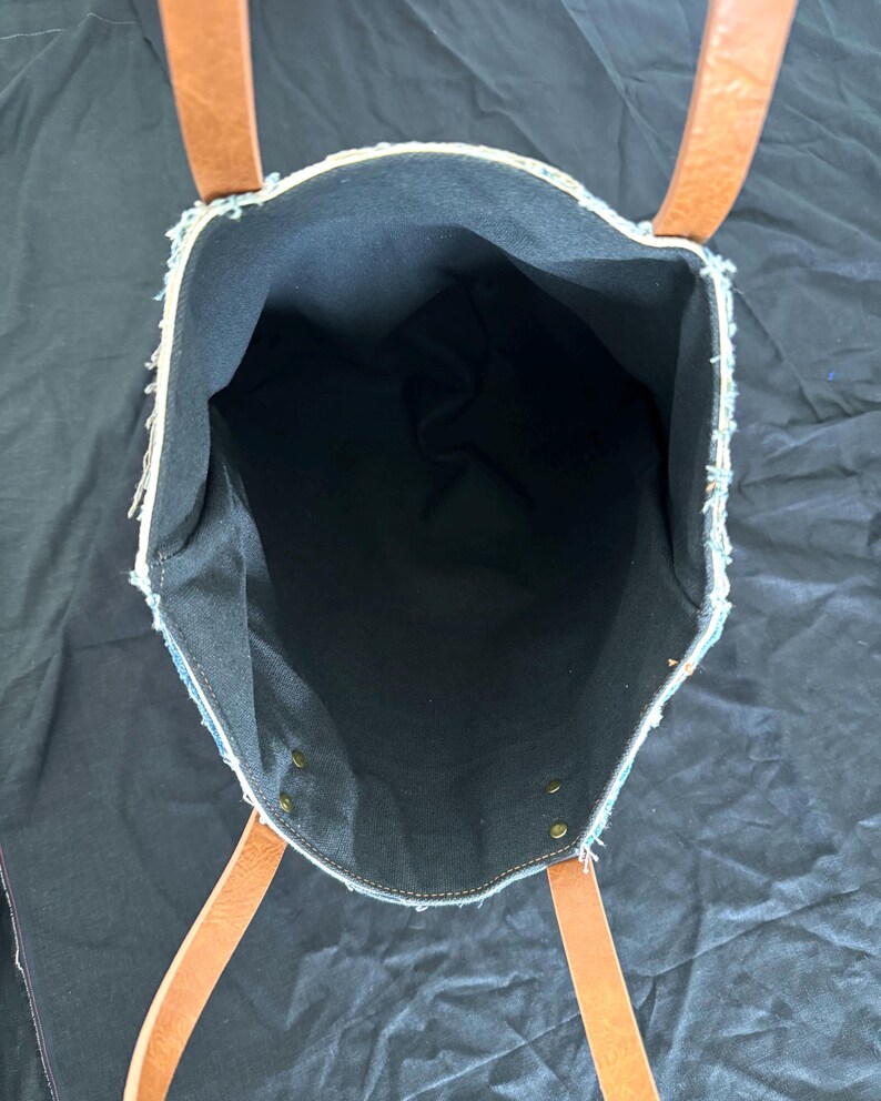 Pu&ograve; includere: Vista aperta di una borsa tote in denim con manici in pelle marrone. L'interno della borsa &egrave; nero e i bordi in denim sono sfilacciati. La borsa ha rivetti in ottone e una costruzione robusta, adatta al trasporto di vari oggetti.