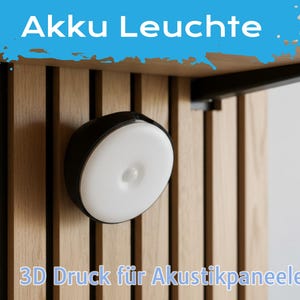Op de afbeelding: Een zwart-witte, cirkelvormige lamp is gemonteerd op een houten paneel. De lamp heeft een witte, cirkelvormige lichtbron. De tekst "Akku Leuchte" staat in een blauwe banner bovenaan en "3D Druck für Akustikpaneele" onderaan.