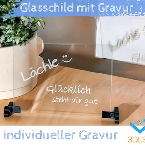 Op de afbeelding: Een helder glazen bord met een witte inscriptie die "Lächle - Glücklich steht dir gut!" luidt. Het bord is gemonteerd op een houten oppervlak met zwarte metalen beugels. Het bord is een gepersonaliseerd gegraveerd glazen bord.