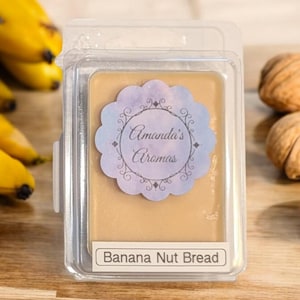 Può includere: Un contenitore di plastica trasparente con un fondente di cera all'interno. Il fondente di cera è di colore marrone chiaro e presenta il testo "Amanda's Aromas" e "Banana Nut Bread".