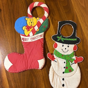 Puede incluir: Dos adornos navideños de tela para colgar en la puerta. Uno es un calcetín rojo con un osito de peluche y la frase "Merry Christmas". El otro es un muñeco de nieve con sombrero negro, bufanda verde y mitones rojos. Ambos tienen un lazo para colgar.