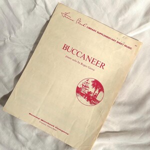 May include: A vintage sheet music cover for a piano solo titled "Buccaneer" by Roger Grove. The cover features a red illustration of a ship sailing on the ocean with palm trees in the background. The text "France Clack Library Supplementary Sheet Music" is printed in red at the top of the cover.