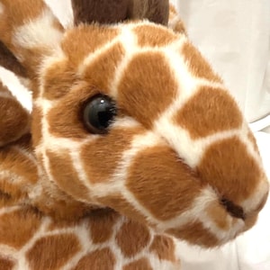 May include: Close-up of a plush giraffe head. The giraffe has a brown and white patterned coat, with dark brown ears and a large, black eye. The soft toy is made of a fuzzy material.