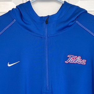 Puede incluir: Una sudadera con capucha azul real con cierre de media cremallera. Presenta la palabra "Tulsa" en escritura roja y un logotipo blanco de Nike. Las costuras blancas acentúan las costuras. La sudadera está colgada en una percha blanca.