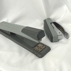 May include: Two gray staplers are displayed on a white surface. One is a larger, rectangular stapler with a gray top and a darker gray base. The other is a smaller, handheld stapler with a silver metal component.