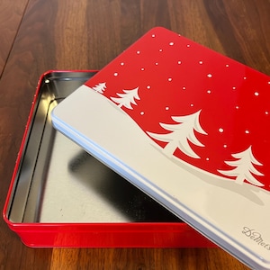 May include: A red rectangular tin with a lid featuring a winter scene. The lid has a red background with white snowflakes and white pine trees. The tin is open, revealing a silver interior. The brand name "DeMet's" is printed on the lid.