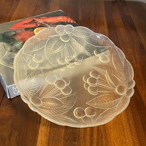 Puede incluir: Un cuenco de vidrio transparente con un diseño de cerezas. El cuenco tiene un borde festoneado y está decorado con cerezas y hojas grabadas. El cuenco está sobre una superficie de madera.