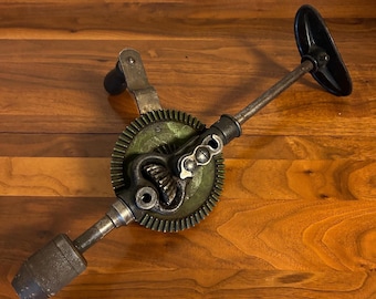 Puede incluir: Un taladro manual antiguo con un mango negro y un mecanismo de engranajes. El taladro tiene un cuerpo de metal con un agarre texturizado y un mango negro. El engranaje es verde y el taladro está sobre una superficie de madera.