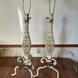 Könnte beinhalten: Paar cremefarbene Tischlampen mit einem Used-Look-Finish. Jede Lampe hat ein geflochtenes, käfigartiges Design auf der Mittelsäule, das von drei gebogenen Beinen getragen wird. Die Lampen haben eine graue Kugel oben und ein braunes Kabel.