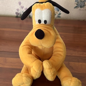 May include: A plush toy of Pluto, a yellow dog with black ears and a black nose. The toy is sitting with its arms and legs extended. The background is a wooden surface.