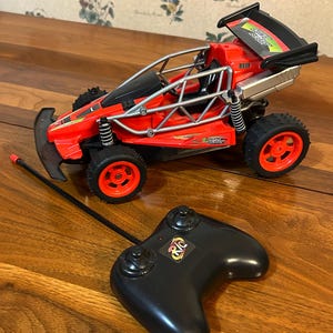 May include: A red and black remote-controlled toy car with black wheels and a silver exhaust. The car has a black spoiler and a black remote control with the letters "RC" on it. The toy car is on a wooden surface.