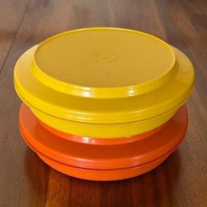 Könnte beinhalten: Zwei Vintage-Kunststoff-Lebensmittelbehälter. Der obere Behälter ist gelb und der untere Behälter ist orange. Die Behälter sind übereinander gestapelt. Die Behälter haben eine runde Form.