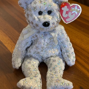 Puede incluir: Un oso de peluche blanco cubierto de lentejuelas plateadas en forma de estrella. El oso tiene ojos negros de botón, una nariz negra y una cinta plateada alrededor del cuello. Una etiqueta en forma de corazón está unida a la oreja.