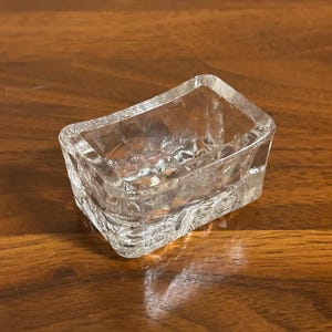 May include: A clear, rectangular glass dish with a textured, ice-like appearance. The dish has a slightly curved shape and is designed with a rough, uneven surface. The glass is transparent, allowing light to pass through, and is likely used for decorative or functional purposes.