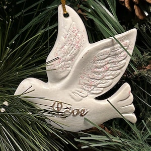 May include: White ceramic dove ornament with pink accents on the wings and the word "Love" in gold script. The dove is hanging from a gold string, set against a backdrop of green pine needles.