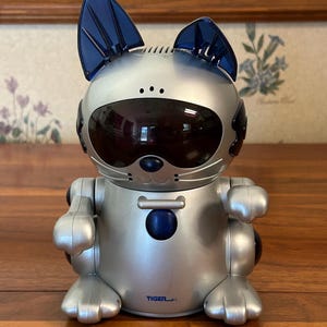 May include: Silver and blue robotic cat toy with a black visor and blue ears. The toy has a round body, cat-like features, and articulated limbs. The word "TIGER" is printed on the front.