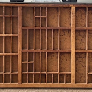 May include: A vintage wooden printer's tray with multiple compartments of varying sizes. The tray is made of light brown wood and features a grid-like design, ideal for organizing small items or displaying collectibles. The tray is likely used for typography.