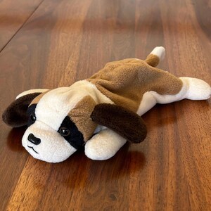 Könnte beinhalten: Ein Stofftier, das einen Bernhardiner-Hund darstellt, liegt auf einer Holzoberfläche. Das Spielzeug hat einen weißen Körper mit braunen Flecken auf Rücken und Kopf sowie dunkelbraune Ohren. Der Hund hat eine schwarze Nase und Augen.