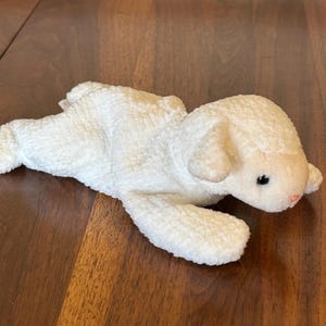 Peut inclure: Un jouet en forme d'agneau en peluche blanche avec un extérieur texturé et duveteux. L'agneau est couché sur une surface en bois. Il a de petits yeux noirs et un nez rose.
