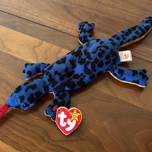 May include: A plush lizard toy featuring a bright blue body with black spots, a long tail, and a red felt tongue. The stuffed animal has a heart-shaped red tag with the white letters ty and a yellow star attached to one of its front legs.