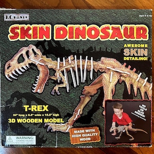 May include: A boxed T-Rex 3D wooden model kit. The box features a detailed illustration of the assembled dinosaur skeleton with "SKIN DINOSAUR" in red letters. The model is 61 cm long, 22 cm wide, and 39 cm high.