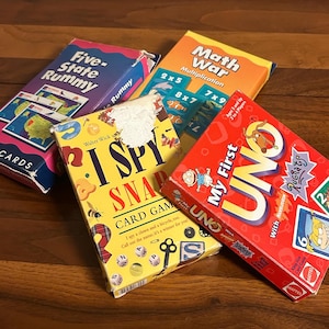 May include: A collection of colorful card game boxes on a wooden surface. Games include "Five-State Rummy," "I Spy Snap," "Math War," and "My First Uno." The boxes feature various designs and text, including the game names and instructions.