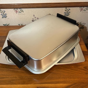 Könnte beinhalten: Ein rechteckiger, silberfarbener Elektrogrill mit schwarzen Griffen. Der Grill steht auf einem passenden silbernen Tablett. Das Bild ist auf einer Holzoberfläche mit Blumengardinen im Hintergrund aufgenommen.
