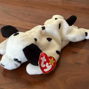 May include: A plush dalmatian dog toy with white fur and black spots, featuring black ears and a black tail. A red heart-shaped tag with the white text 'ty' and the gold text 'BEANIE ORIGINAL BABY' is attached to the front of the plush toy.