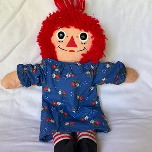 May include: A vintage Raggedy Ann doll with a red yarn hair, a painted face, and a blue floral dress. The doll has striped red and white socks and black shoes. A classic children's toy.