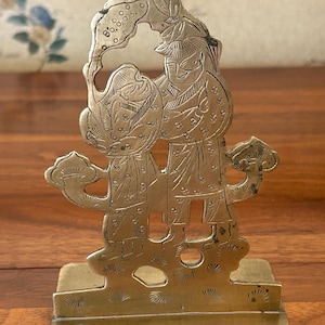 Peut inclure: Figurine en métal couleur laiton représentant deux personnages en tenue asiatique traditionnelle. La gravure détaillée montre les personnages avec des chapeaux et des vêtements ornés, sur une base rectangulaire. Le design comprend des motifs floraux et nuageux.