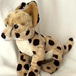 Puede incluir: Un peluche de guepardo con cuerpo beige, manchas marrones oscuras y detalles negros en las orejas y la melena. El animal de peluche está sentado, con la cabeza girada hacia la izquierda.