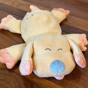 Puede incluir: Un juguete de peluche con forma de perro en posición tumbada. El juguete es principalmente amarillo con detalles melocotón en las patas, orejas y nariz. Tiene una nariz azul y ojos bordados. El juguete está hecho de tela suave y texturizada.