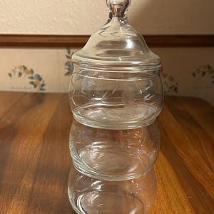 Pode incluir: Um frasco de vidro transparente de três camadas com tampa e alça. O frasco tem desenhos florais gravados e é empilhado um em cima do outro. O frasco está sobre uma superfície de madeira.