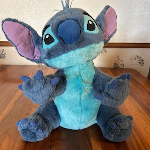 Puede incluir: Un peluche de Stitch, con una combinación de colores azul y turquesa, grandes orejas y ojos expresivos. El muñeco tiene un mechón de pelo en la cabeza y posa con los brazos y las piernas extendidos. Un personaje clásico de Disney.