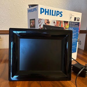 Puede incluir: Un marco de fotos digital negro con un acabado brillante. El marco es rectangular y tiene una pantalla negra. Una caja con la marca "PHILIPS" está detrás del marco. La caja muestra imágenes de personas y el texto "8" y "Digital PhotoFrame".