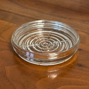 May include: A clear glass coaster with a circular design. The coaster has concentric rings and a central pattern. The glass is transparent, allowing the surface beneath to be visible. The coaster is designed to protect surfaces from drink condensation.