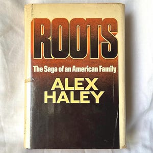 May include: A vintage hardcover book titled "ROOTS" by Alex Haley. The cover features the title in large orange and brown letters, with the subtitle "The Saga of an American Family" below. The author's name is in yellow letters. The background is a gradient of brown and tan.