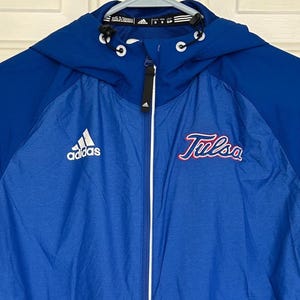 Puede incluir: Chaqueta con capucha Adidas azul con cremallera completa. La chaqueta presenta el logotipo de Adidas en el pecho izquierdo y la palabra "Tulsa" en escritura roja en el pecho derecho. La chaqueta tiene capucha y cremallera blanca.