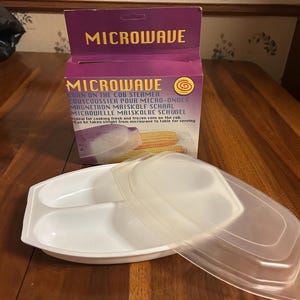 Puede incluir: Un vaporizador de maíz para microondas blanco con tapa transparente y una caja morada y blanca. La caja dice "MICROWAVE" y enumera el nombre del producto en varios idiomas. El vaporizador está diseñado para cocinar maíz.