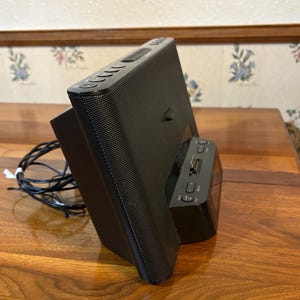 May include: Black digital clock radio with a speaker and a docking station. The clock has a rectangular shape with several buttons on top and a small display. The speaker has a mesh front and is angled slightly.