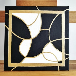 May include: Abstract painting on canvas with a black background and white geometric shapes outlined in gold. The painting is framed with a gold border.
