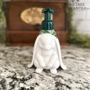 May include: White bunny-shaped soap dispenser with a green pump. The dispenser features a detailed bunny figure at the base, with long ears and closed eyes. The dispenser is on a speckled countertop. The text "LONETREE MERCANTILE" is visible.