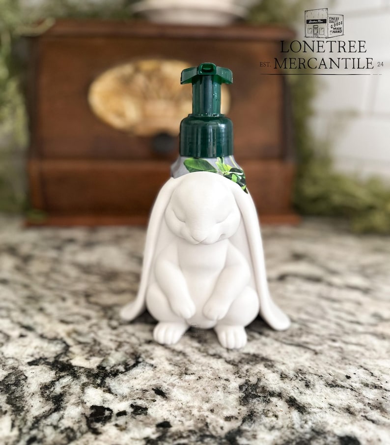 May include: White bunny-shaped soap dispenser with a green pump. The dispenser features a detailed bunny figure at the base, with long ears and closed eyes. The dispenser is on a speckled countertop. The text "LONETREE MERCANTILE" is visible.