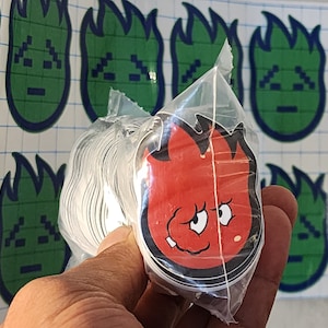 Peut inclure: Gros plan sur une main tenant une pile d'autocollants rouges et noirs. Les autocollants représentent un personnage de dessin animé avec une tête en forme de flamme et une expression espiègle. Les autocollants sont emballés dans un sac en plastique transparent.