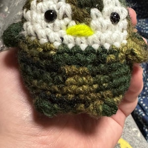 Puede incluir: Un búho de ganchillo hecho a mano en colores de camuflaje. El búho tiene detalles blancos alrededor de los ojos, ojos de cuentas negras y un pico amarillo. El cuerpo es de hilo verde, marrón y beige.