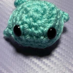 Puede incluir: Una pequeña tortuga de crochet azul turquesa con ojos de botón negros.